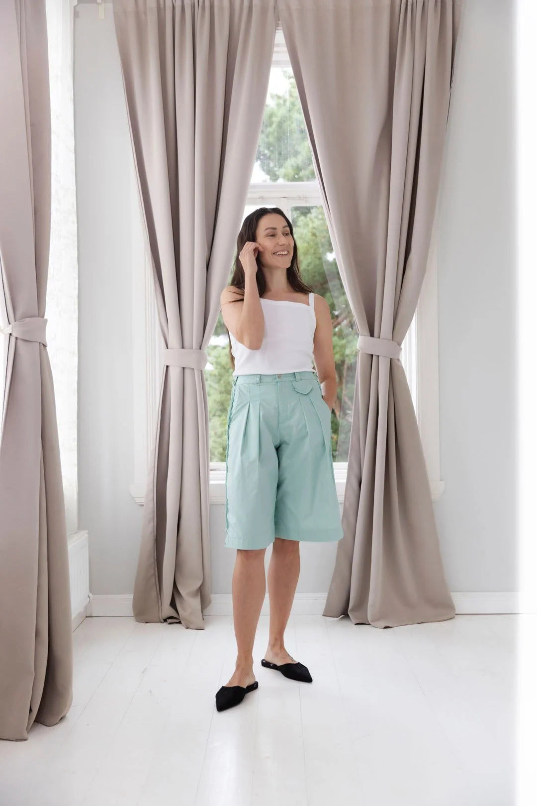 Woman standing in a bright room with large windows and beige curtains.