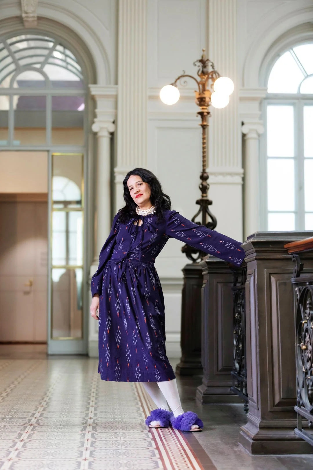 Woman in a purple dress standing in an elegant interior setting with large windows and decorative columns.