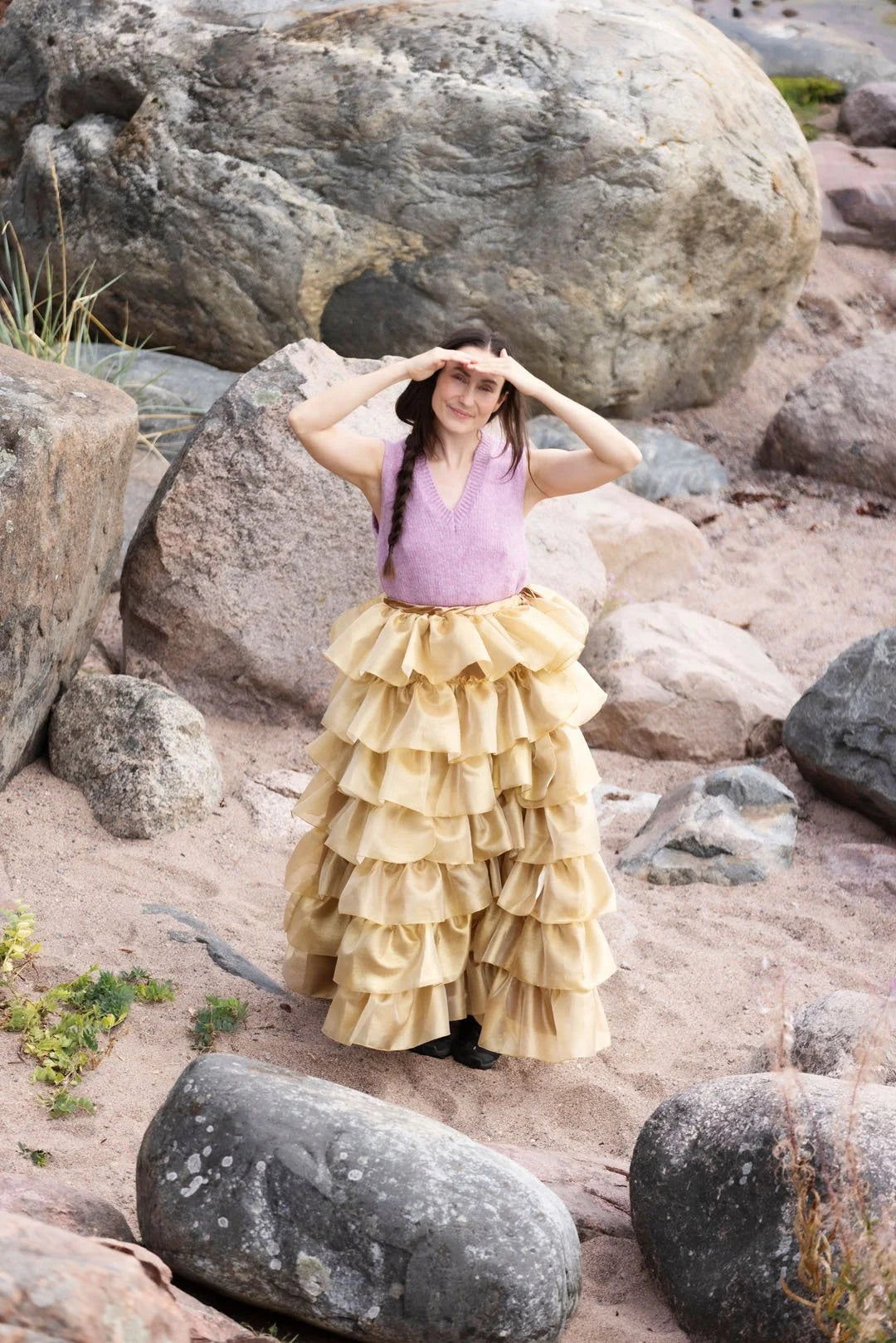 Woman in a yellow ruffled skirt and pink top standing on a rocky beach.