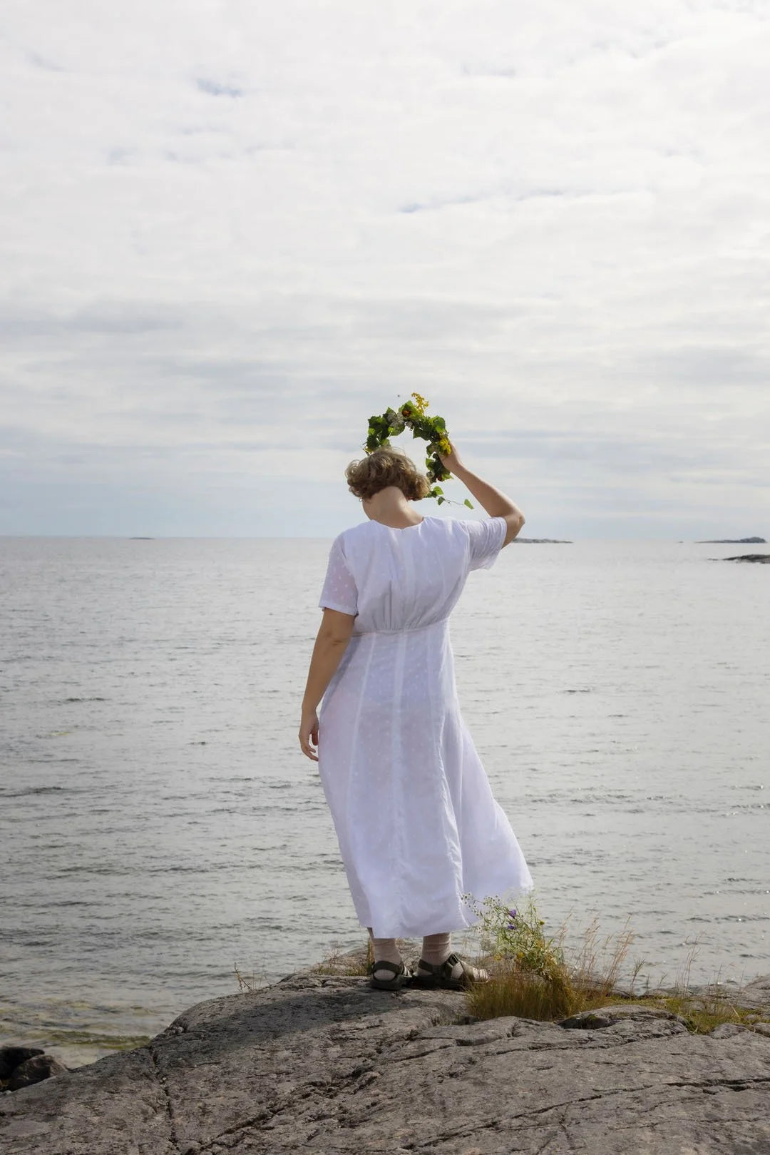 Person in a white dress holding a floral wreath by a body of water