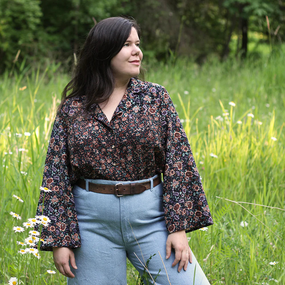 Woman in a floral blouse and light blue pants standing in a grassy field with wildflowers.
