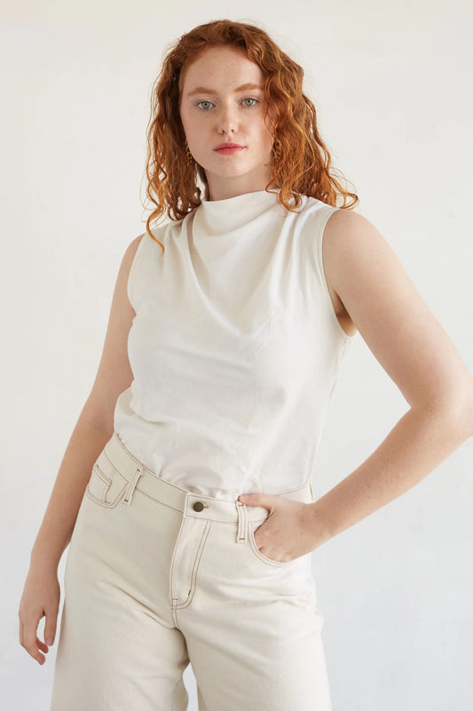 Woman wearing a white sleeveless top and beige pants against a plain background