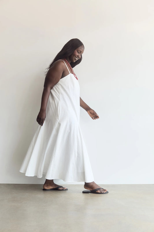 A woman wearing a white sleeveless maxi dress with thin straps and dark flip-flop sandals, standing in a minimalist room with a white wall and light concrete floor.