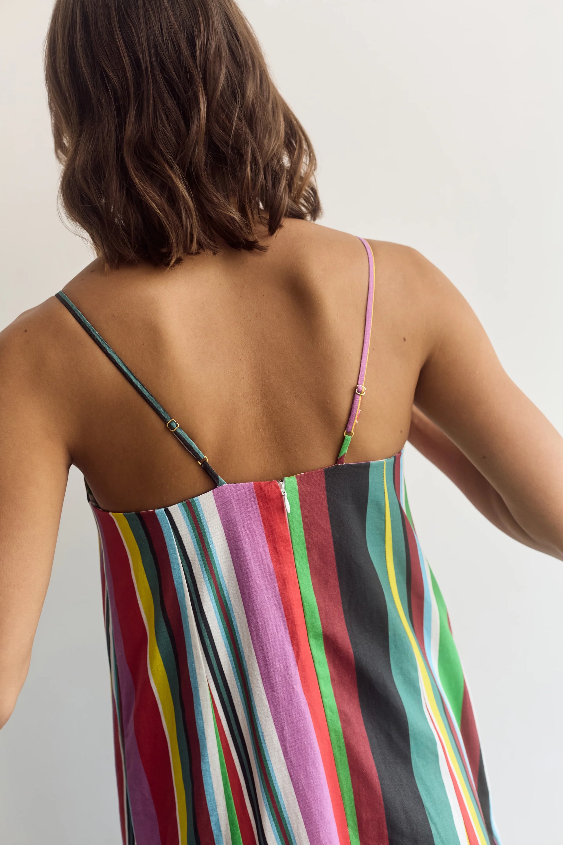 A woman shown from the back wearing a colorful striped camisole dress with thin straps and a multicolor vertical stripe pattern.