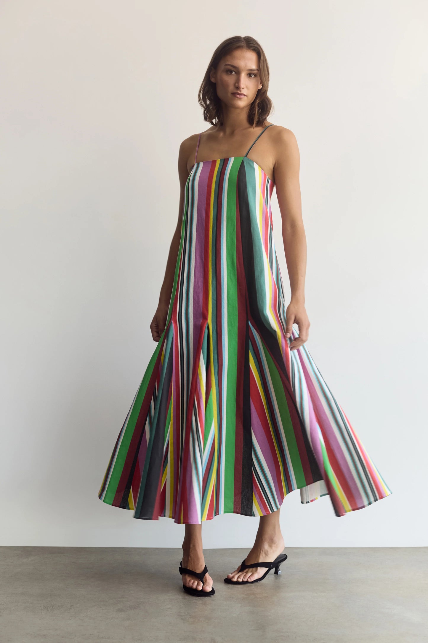 A woman modeling a sleeveless midi dress with vertical multicolor stripes in green, pink, blue, yellow, and white, paired with black heeled sandals against a plain light gray wall.