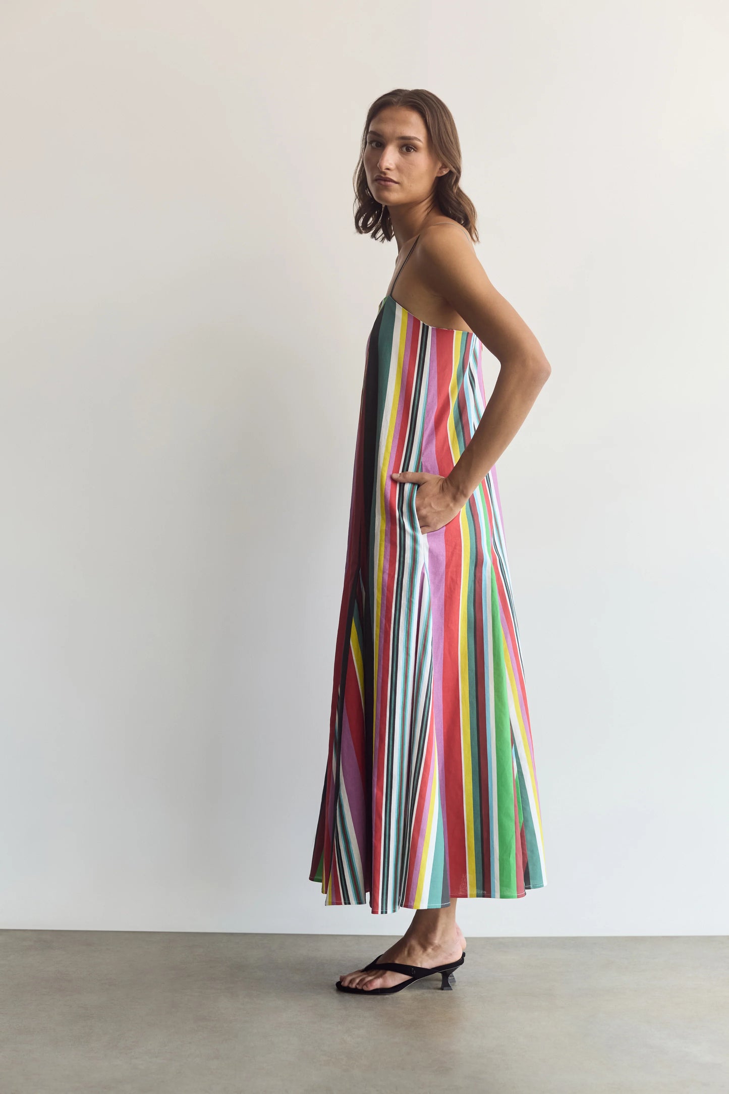 A woman modeling a sleeveless maxi dress with vibrant vertical stripes in pink, green, yellow, blue, and white, standing against a plain white wall.