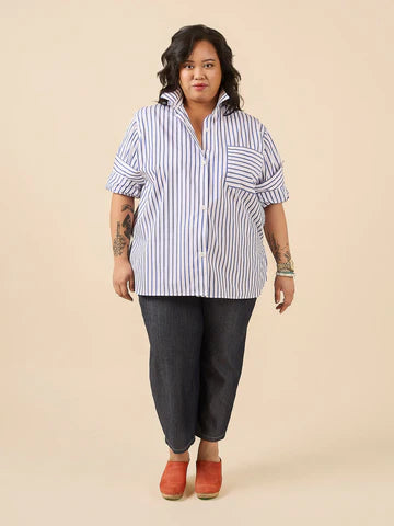 A woman modeling a blue and white vertically striped short-sleeve button-up shirt, paired with dark gray jeans and rust-orange clogs, against a solid beige studio background.