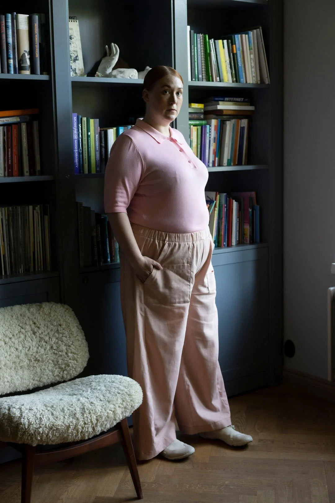 Woman in pink outfit standing in a room with bookshelves and a chair.