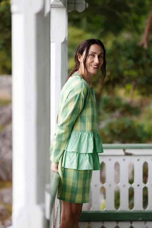Woman in a green checkered dress standing on a porch with a blurred natural background