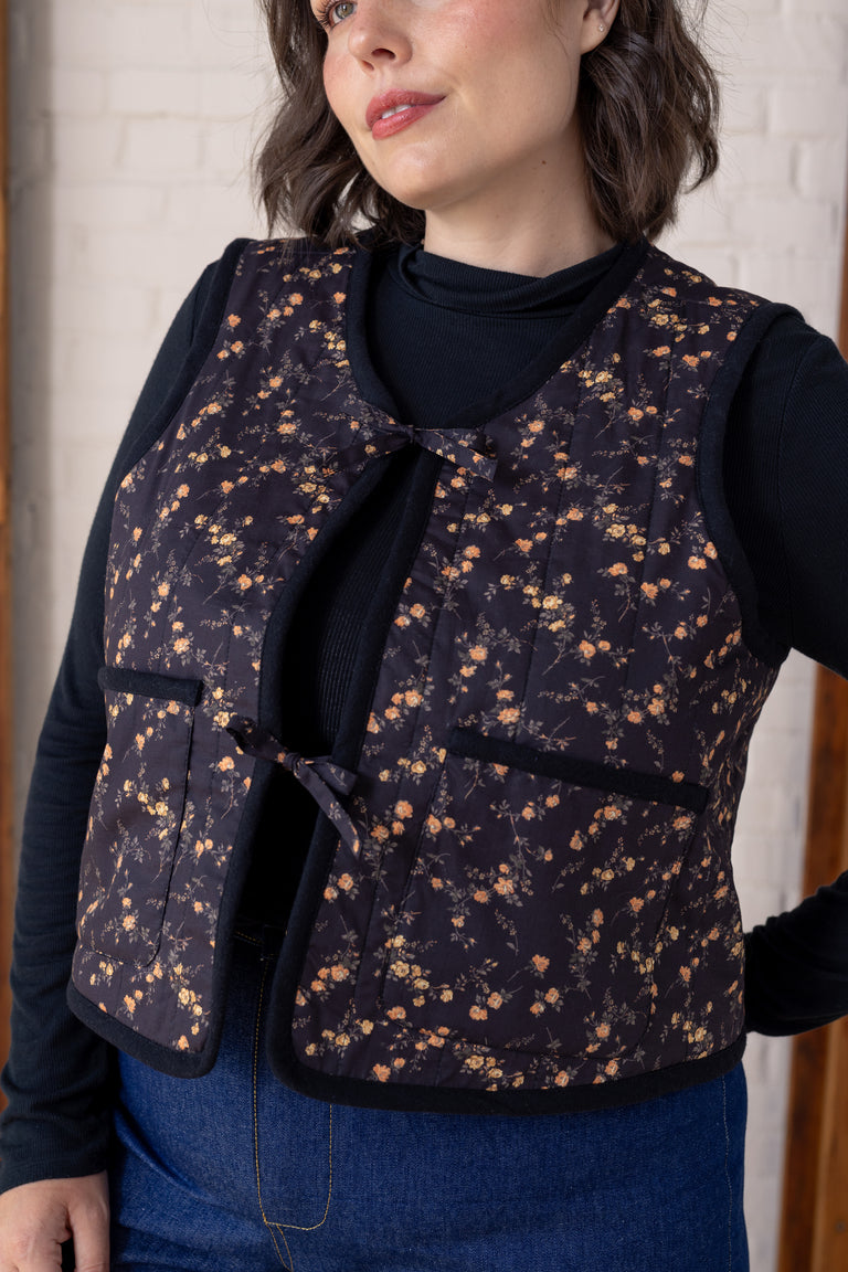 A woman wearing a dark floral quilted vest over a black long-sleeve top and blue jeans, standing indoors against a light brick wall.