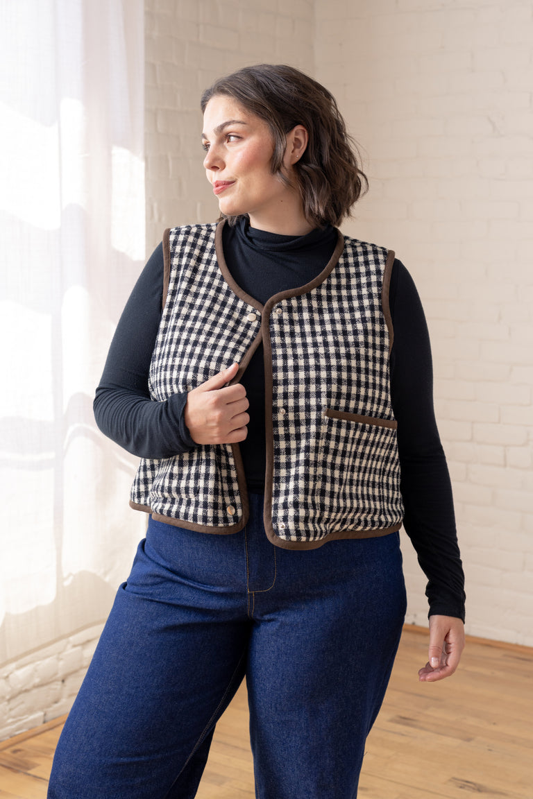 A woman wearing a black and white houndstooth quilted vest with brown trim over a black long-sleeve top and blue jeans, standing in a bright room with a white brick wall and wooden floor.