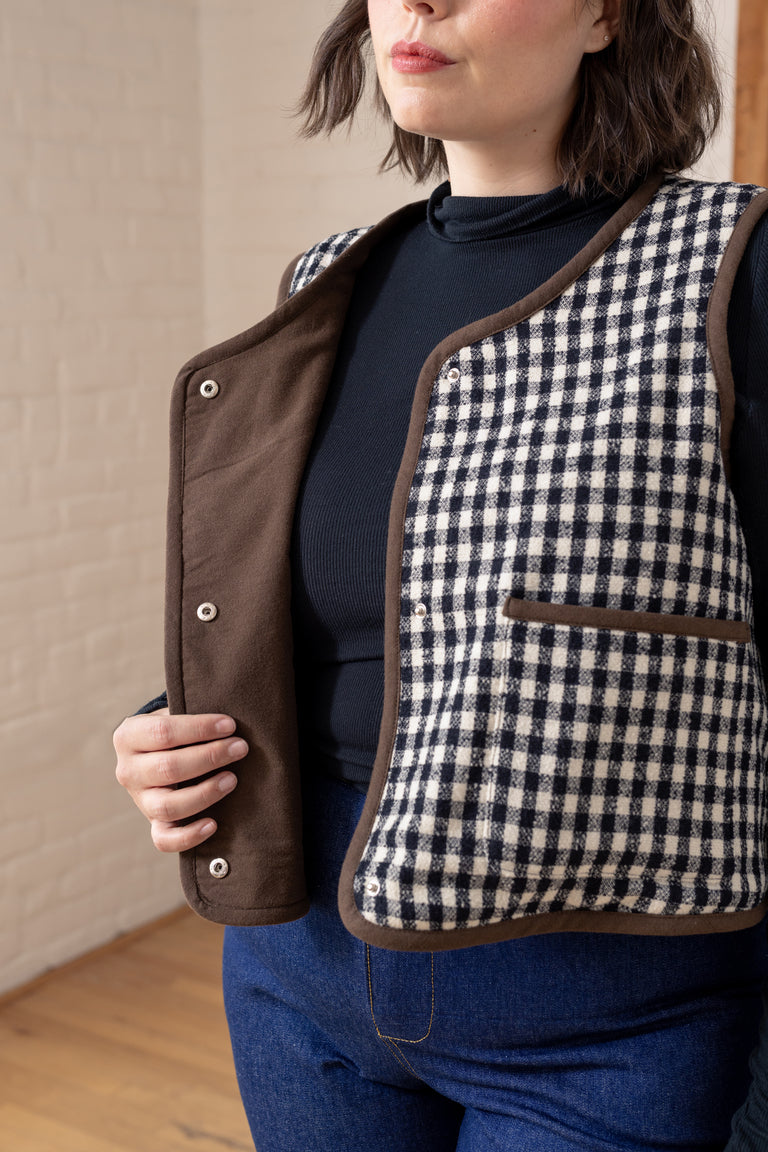 A person wearing a sleeveless quilted vest with a black and white gingham patterned front and brown trim, shown indoors against a light brick wall and wood floor.