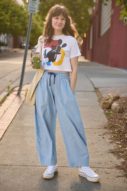 Woman standing on a sidewalk holding a drink, wearing a colorful graphic t-shirt and light blue pants.