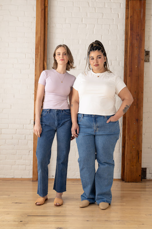 Two women standing side by side wearing matching outfits with blue jeans and white tops against a white brick wall.