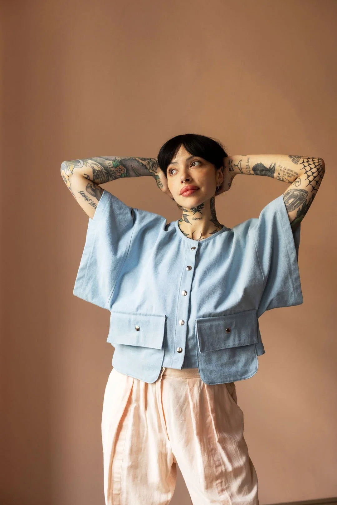 Person wearing a light blue top and pink pants with tattoos on arms against a brown background