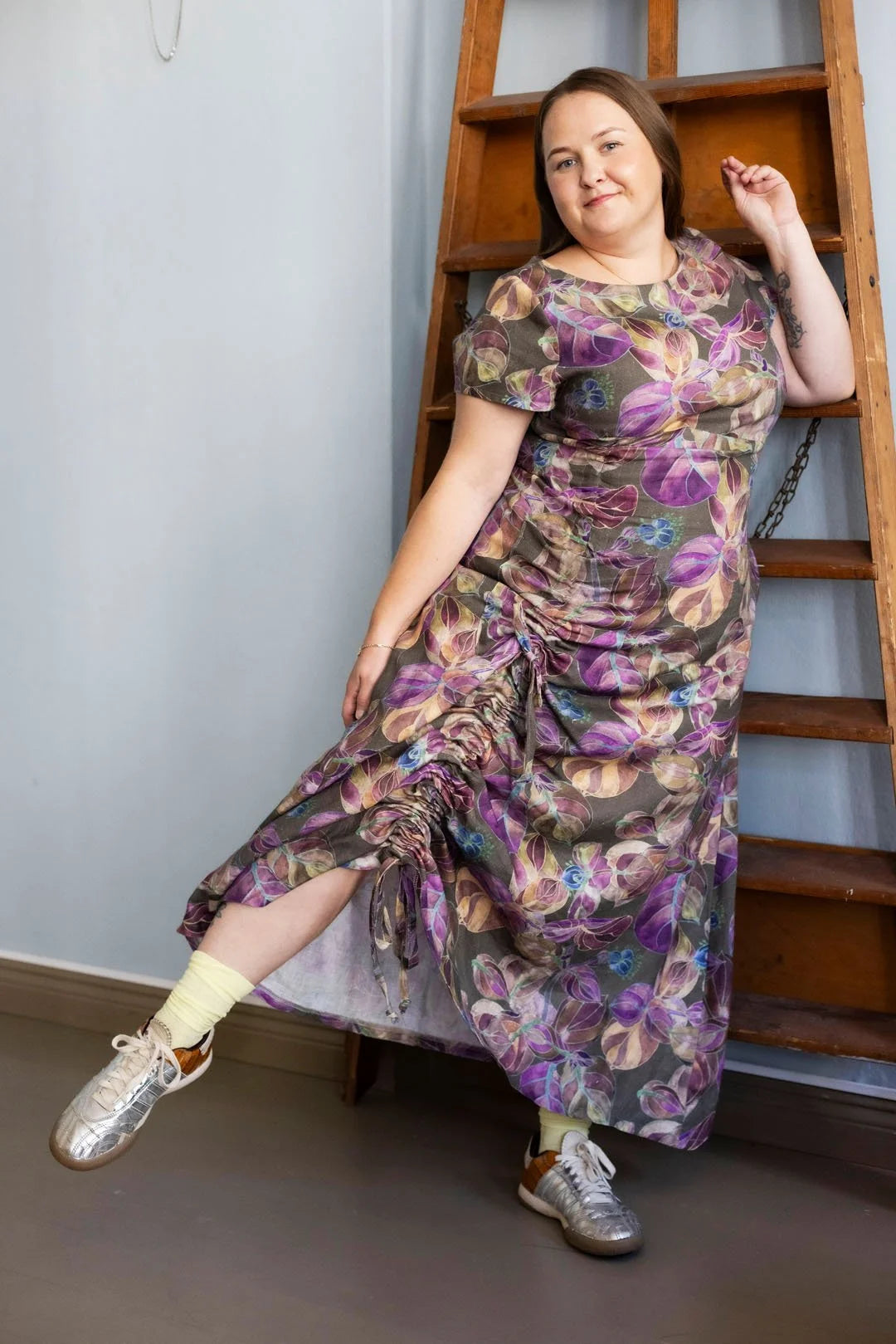 Woman in a floral dress standing next to a wooden ladder indoors.