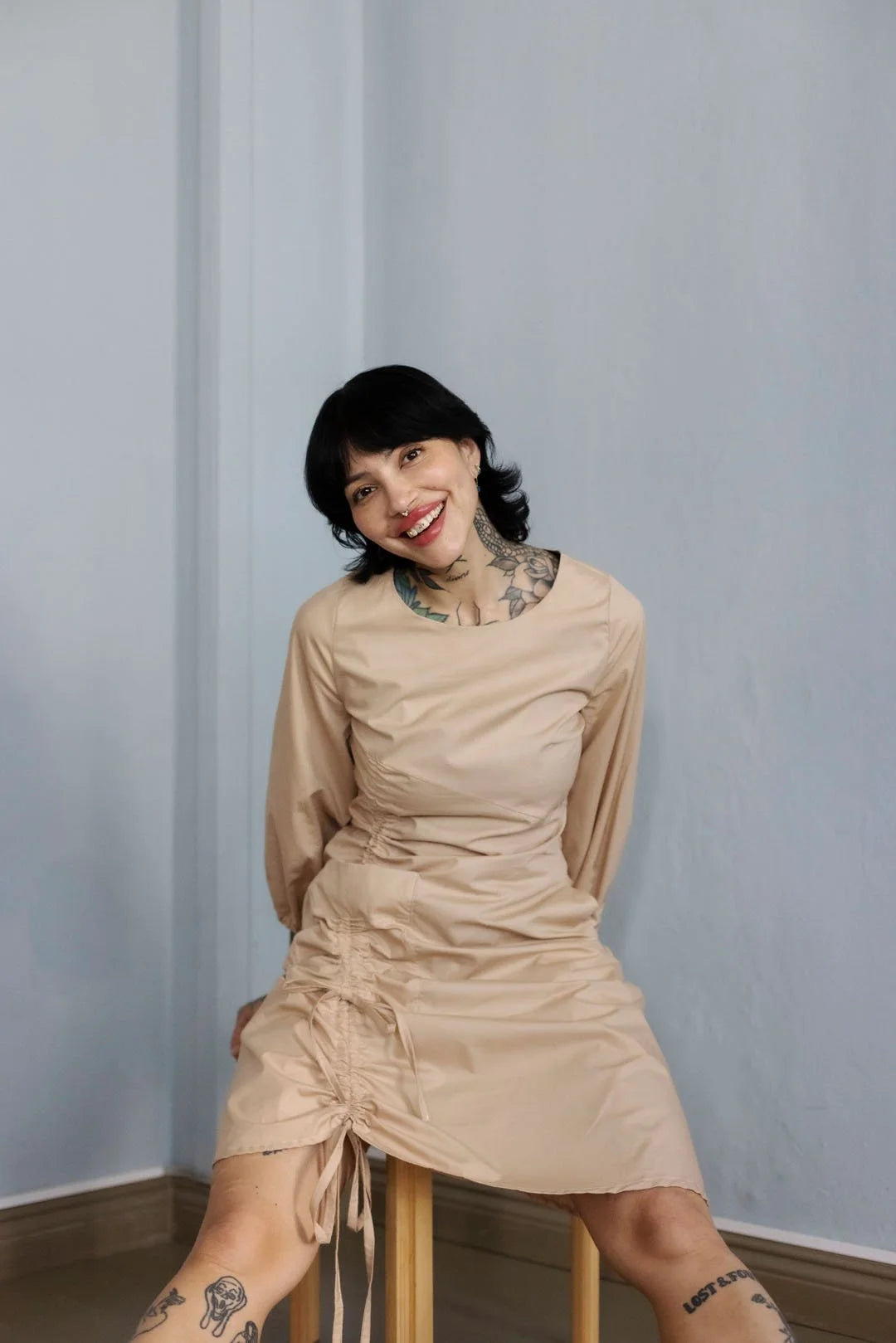 Woman in a beige dress sitting on a stool against a light blue wall.