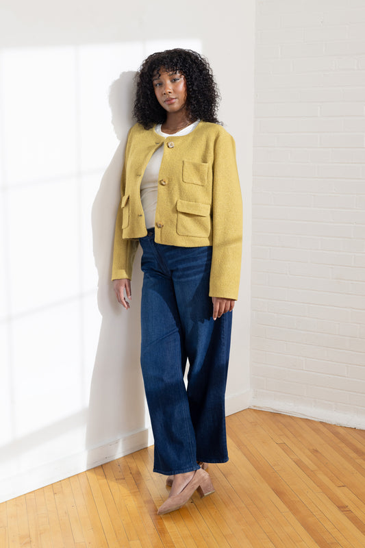 Woman wearing a yellow jacket and blue jeans standing against a white wall.