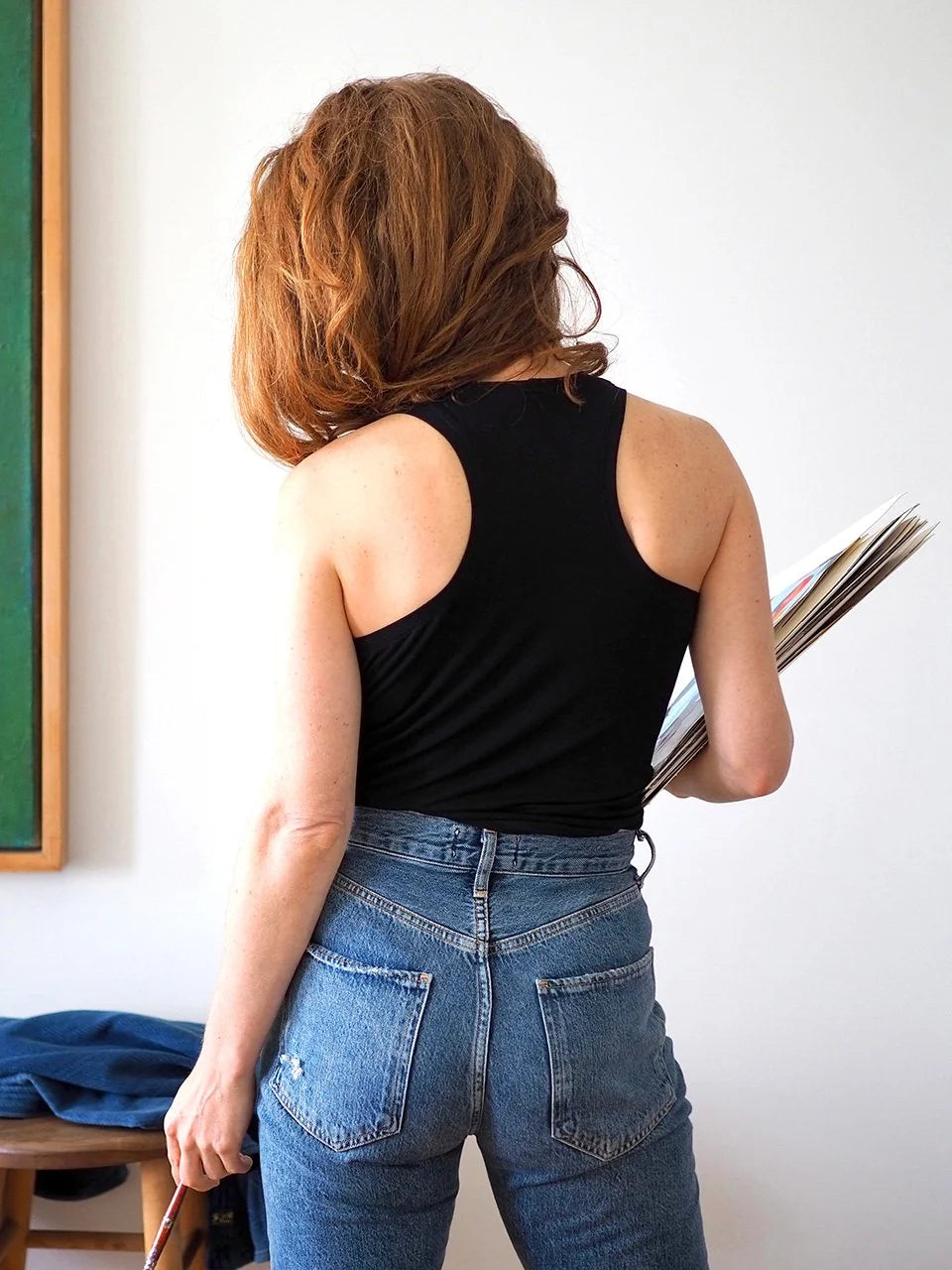 Person wearing a black sleeveless top and blue jeans holding a magazine against a white wall.