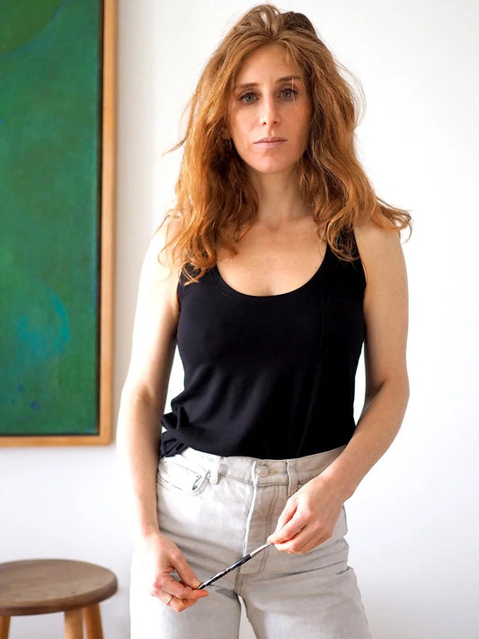 Woman wearing a black tank top and light gray pants standing indoors with a painting in the background.