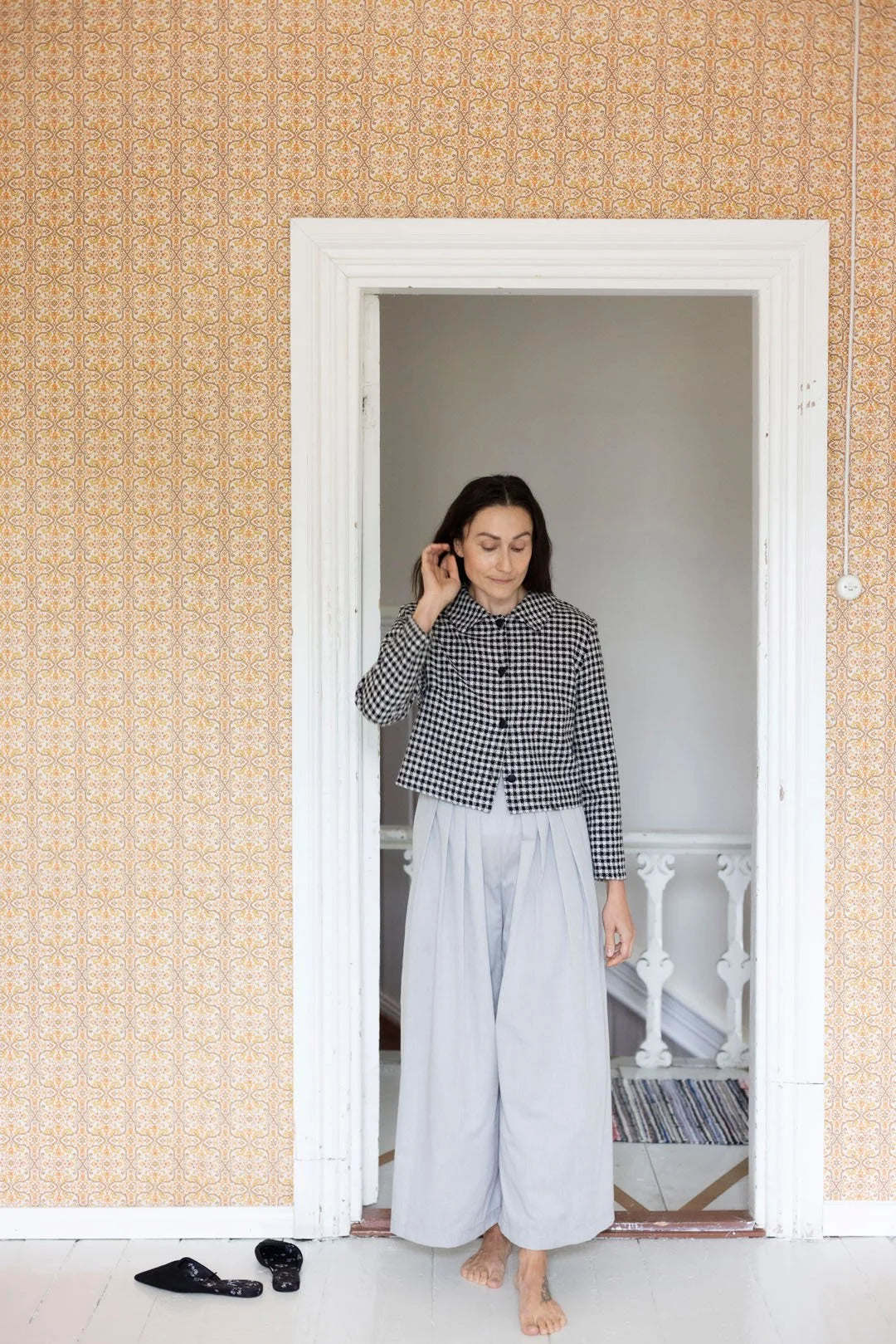 Woman standing in a doorway wearing a checkered shirt and white pants, with a patterned wall behind her.