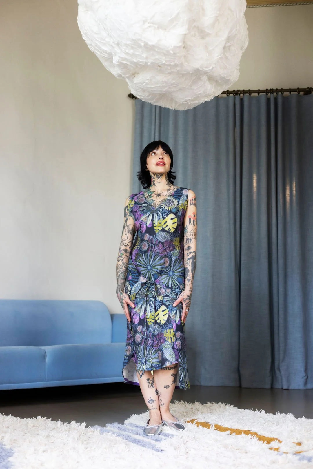 Woman in a floral dress standing in a room with a blue couch and gray curtains.
