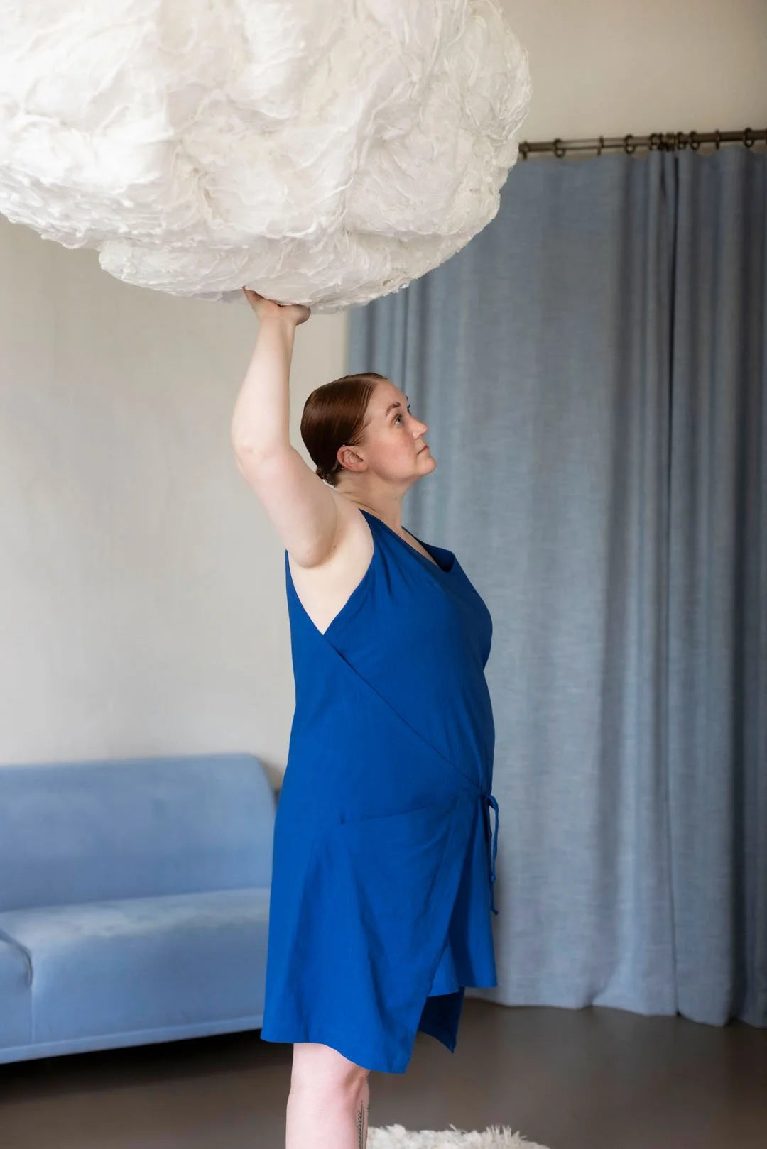 Woman in a blue dress holding a large white cloud-like object in a room with a blue couch and gray curtain.