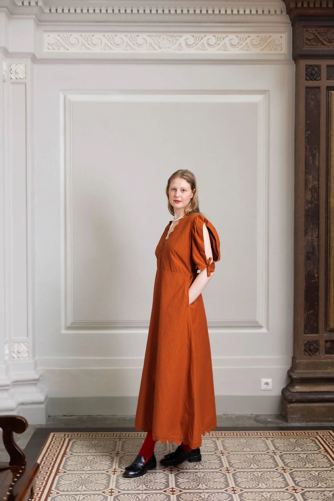 Woman in a rust-colored dress standing in an elegant room with white walls and decorative moldings.