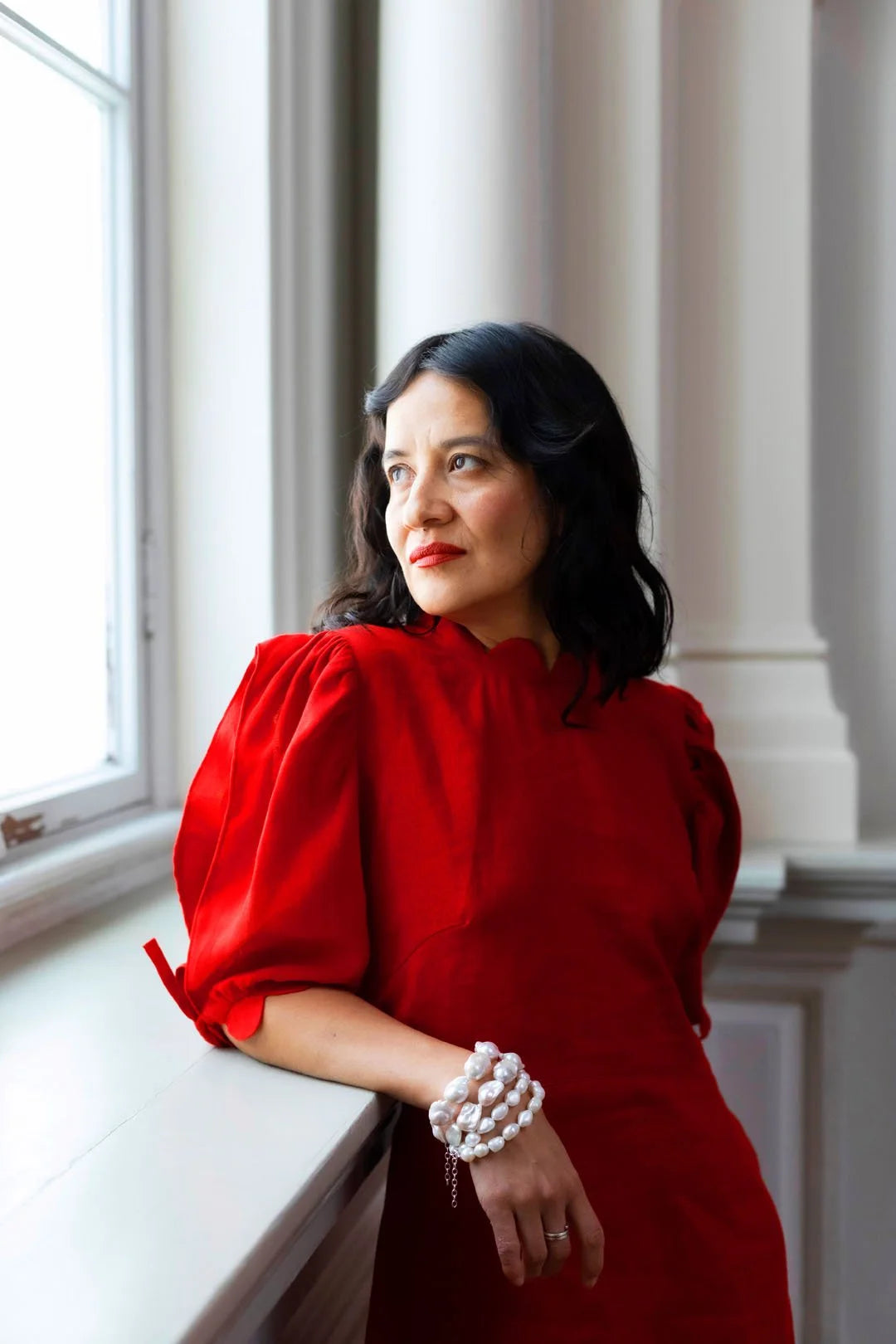 Woman in a red dress standing by a window in an elegant interior setting