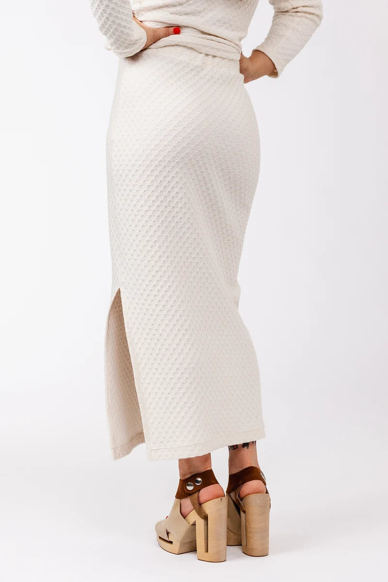 A person shown from the back wearing a textured cream midi skirt with a side slit, paired with brown platform sandals, against a plain white studio background.