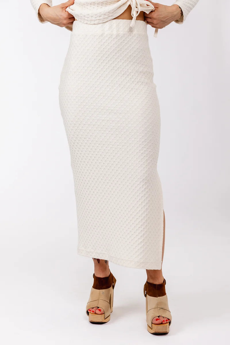 An off-white textured knit midi skirt with a side slit, shown on a model wearing beige wedge sandals against a white studio background.