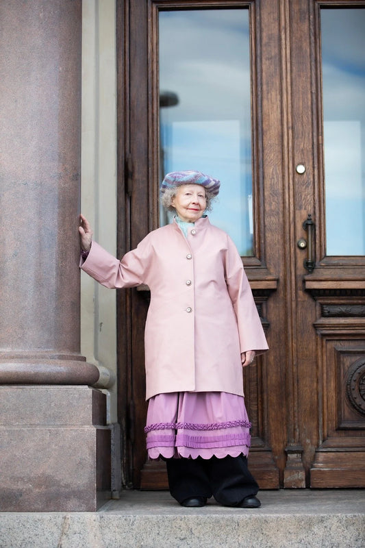 Person in a pink coat and purple hat standing in front of a wooden door.