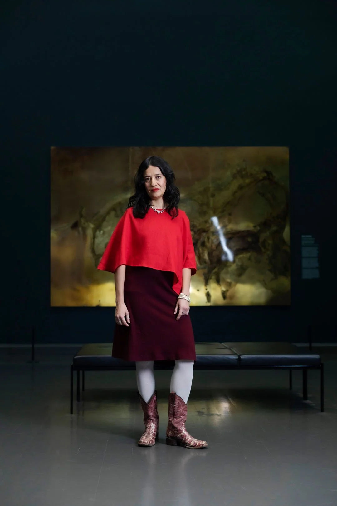 Woman in a red top and maroon skirt standing in front of a large painting in a dark room.