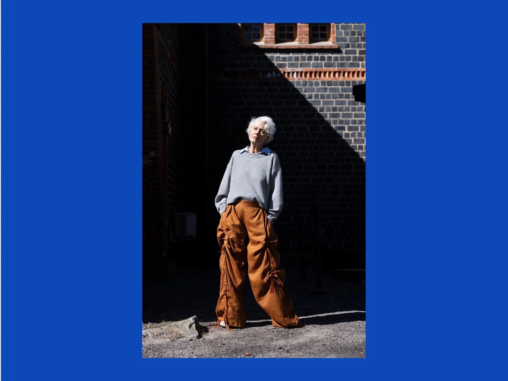 A person standing outdoors in front of a dark brick wall, wearing a light gray long-sleeve top and loose brown wide-leg pants. The photo is centered within a solid blue border.