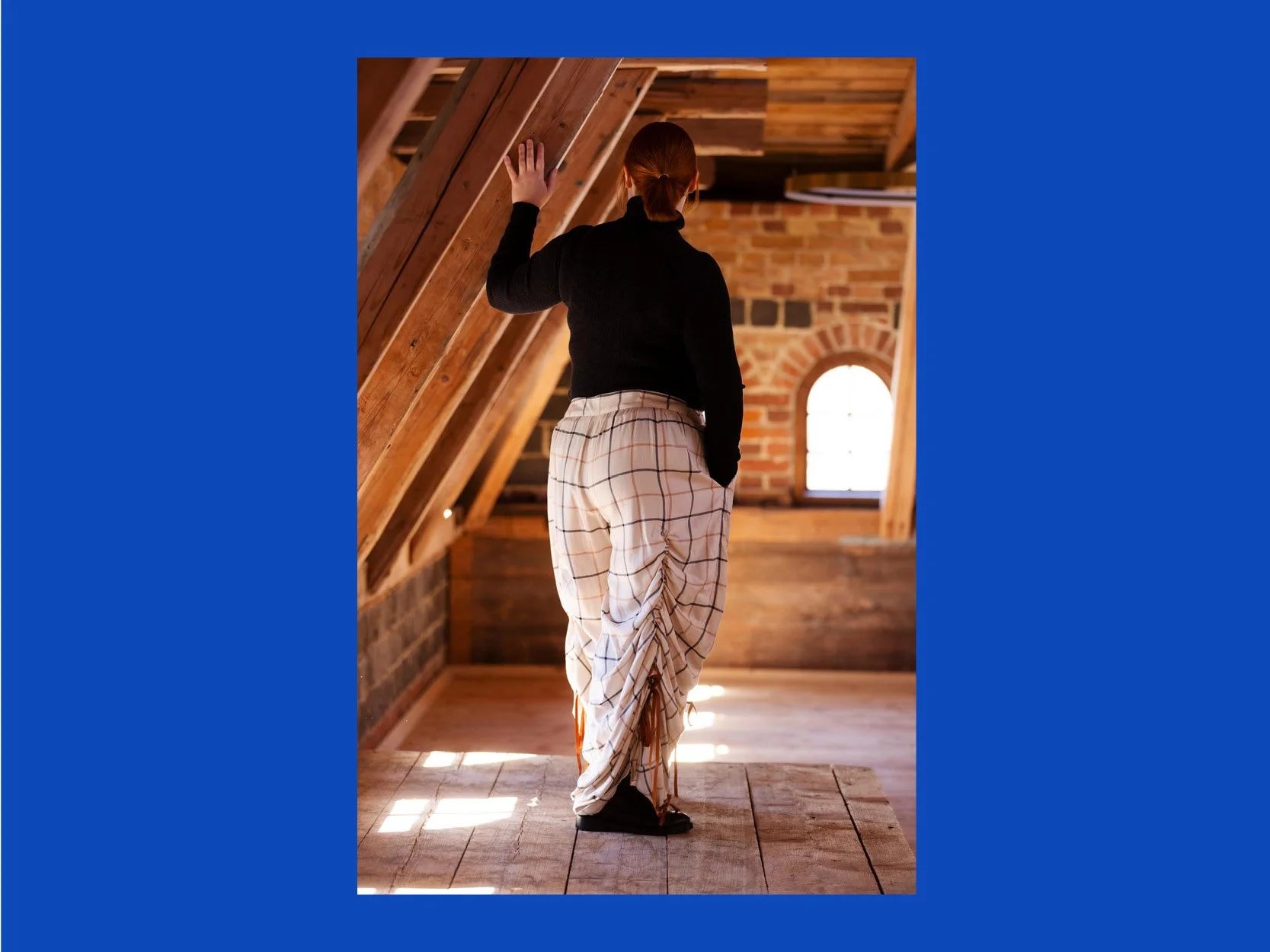 A person standing in a rustic attic or loft space with exposed wooden beams and brick walls, viewed from behind. The person is wearing a black long-sleeve top and loose white pants with a brown windowpane plaid pattern.