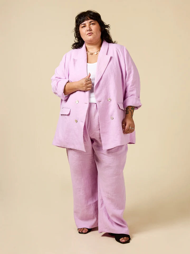 A model wearing a light pink double-breasted linen-blend blazer and matching wide-leg trousers, styled with a white top and black sandals, against a solid beige studio background.