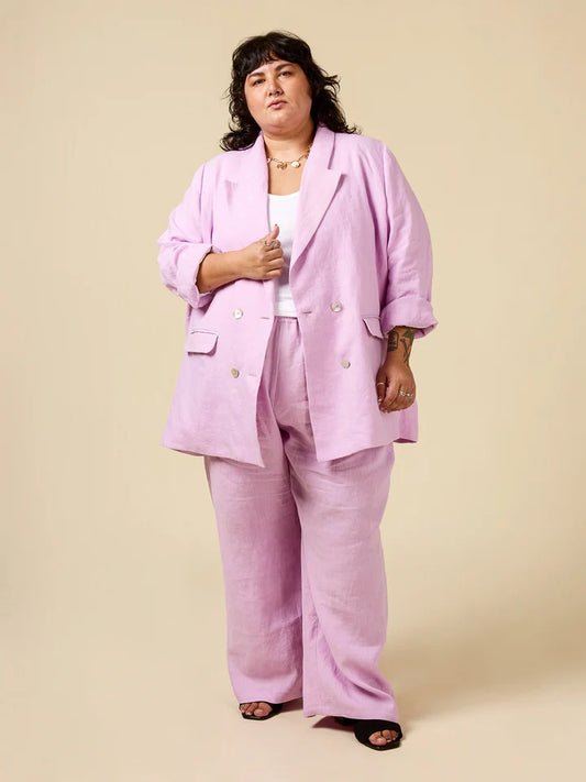 A model wearing a light pink double-breasted linen-blend blazer and matching wide-leg trousers, styled with a white top and black sandals, against a solid beige studio background.