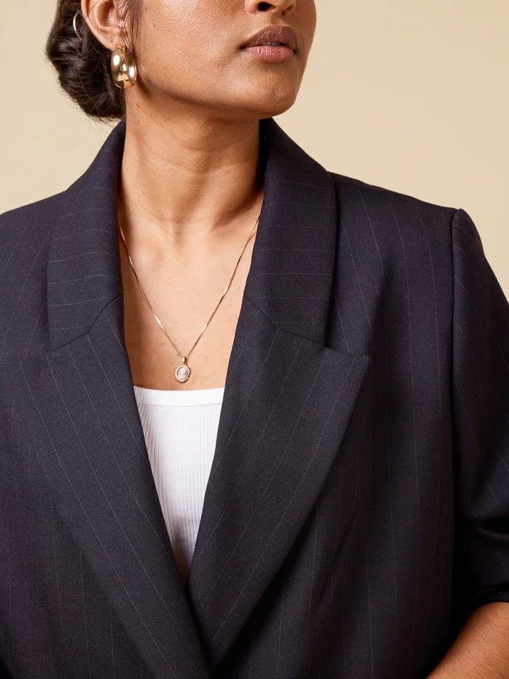 A close-up of a model wearing a dark navy pinstripe blazer over a white top, accessorized with a delicate gold pendant necklace and gold hoop earrings, set against a solid beige background.