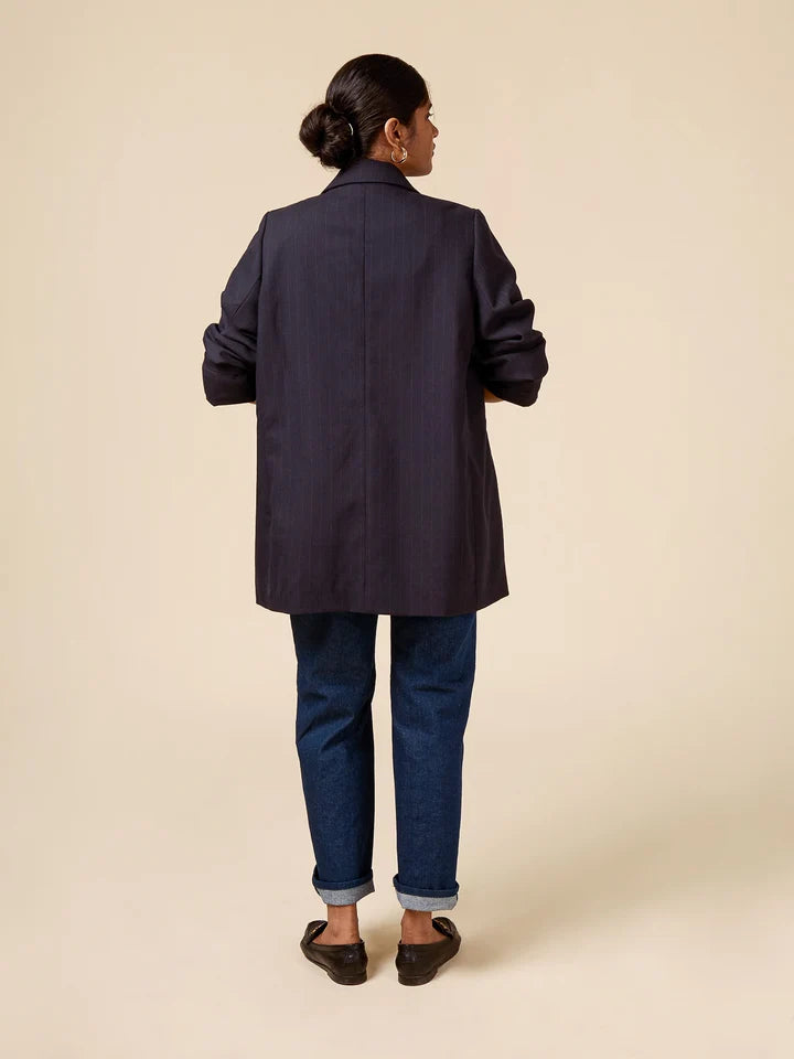A woman shown from the back wearing a dark navy blue blazer, blue jeans, and dark brown shoes against a solid beige studio background.