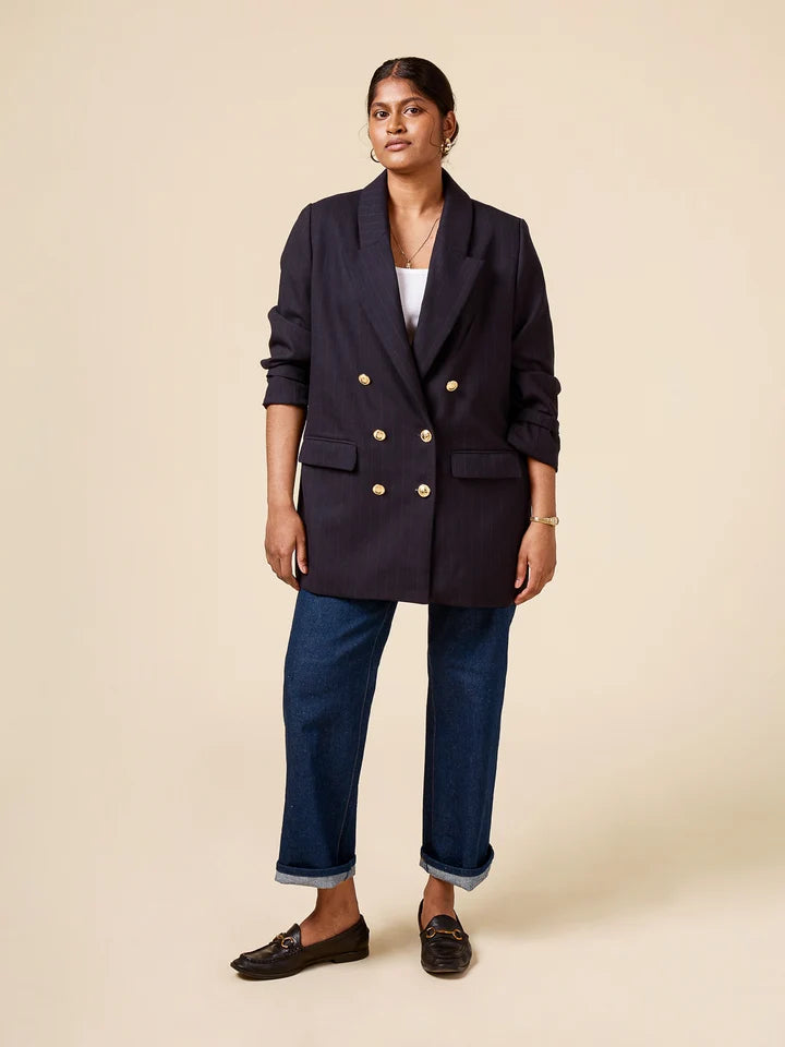 A woman modeling a double-breasted navy blazer with gold buttons, paired with blue jeans and dark loafers, standing against a solid beige studio background.
