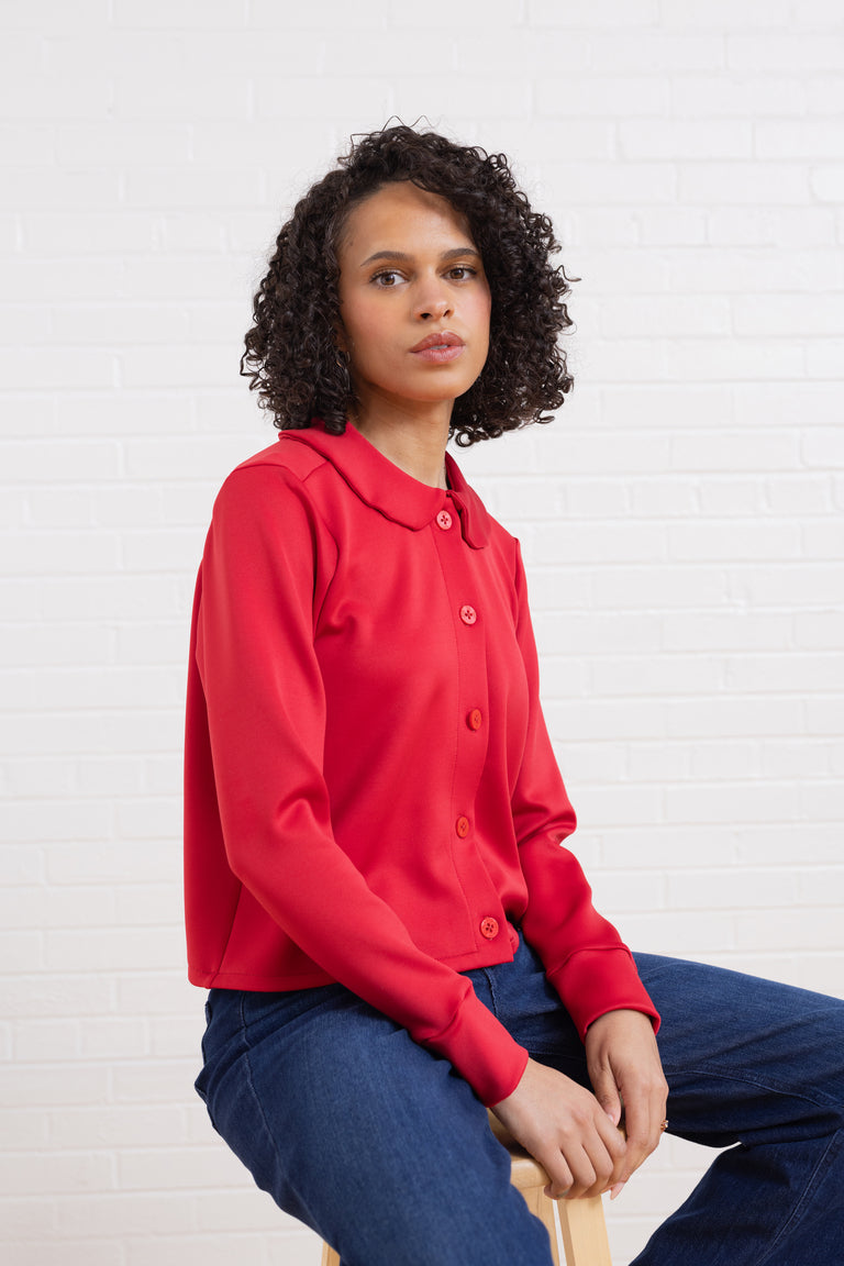 Woman wearing a red shirt sitting on a chair against a white brick wall.