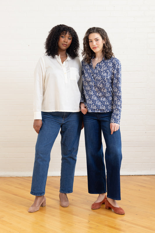 Two women standing side by side wearing jeans and tops against a white wall.