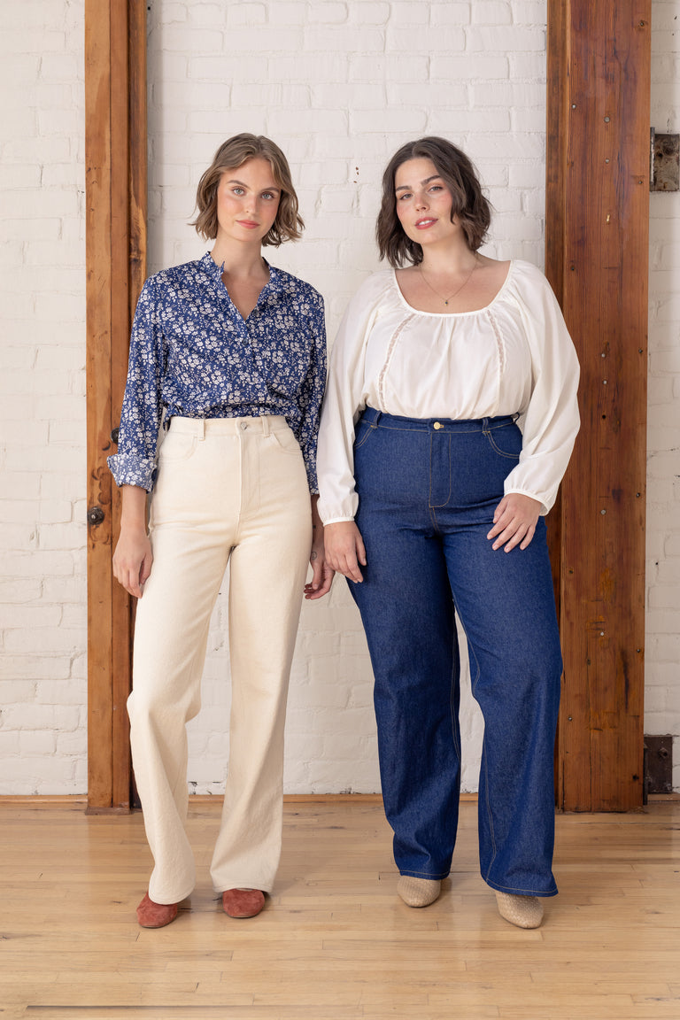 Two women standing indoors against a white brick wall and wooden doorframes, wearing casual outfits. The woman on the left wears a blue floral long-sleeve blouse with cream wide-leg pants.