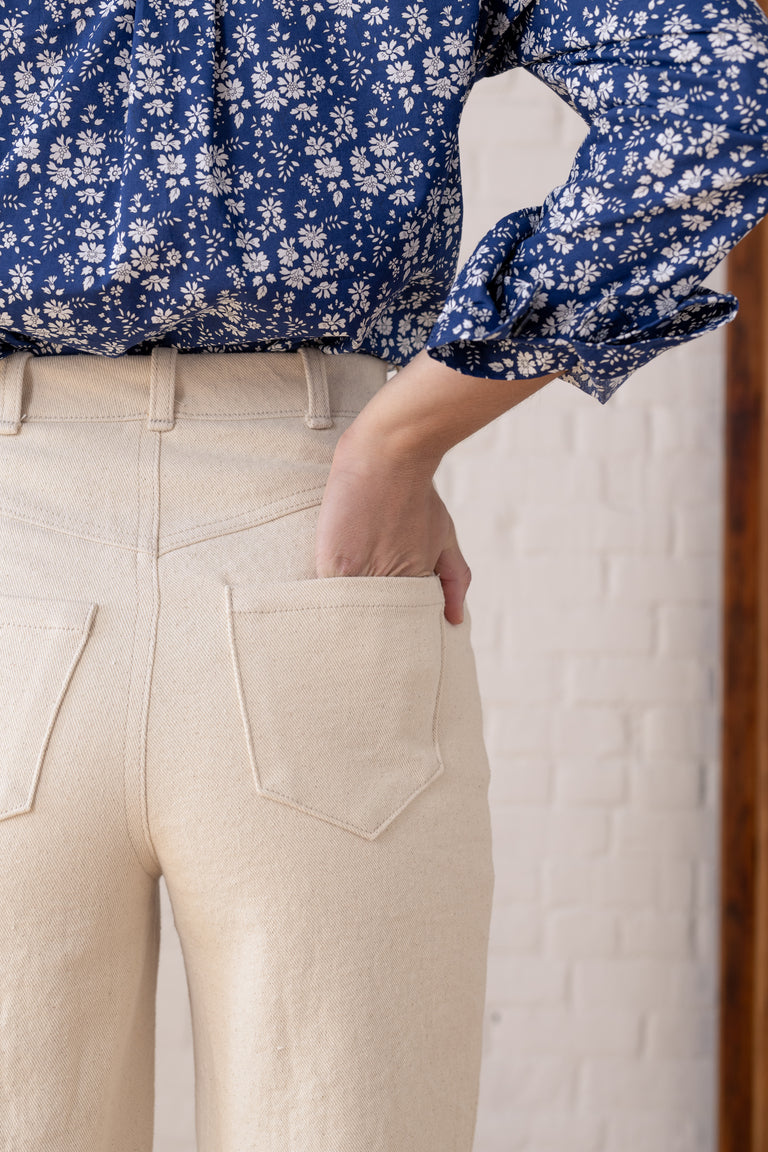 Close-up of a person wearing light beige high-waisted trousers with a back pocket and a blue floral long-sleeve blouse, standing against a white brick wall.
