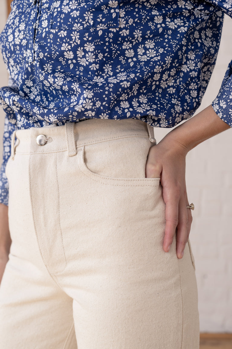 Close-up of a person wearing cream-colored high-waisted linen pants with a blue and white floral blouse, standing against a white brick wall.