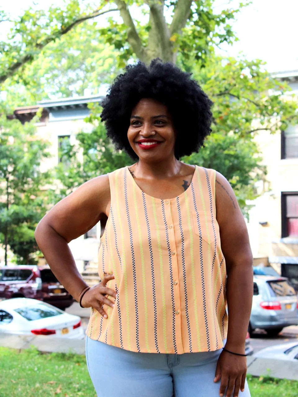 Woman wearing a yellow striped sleeveless top outdoors with trees and buildings in the background