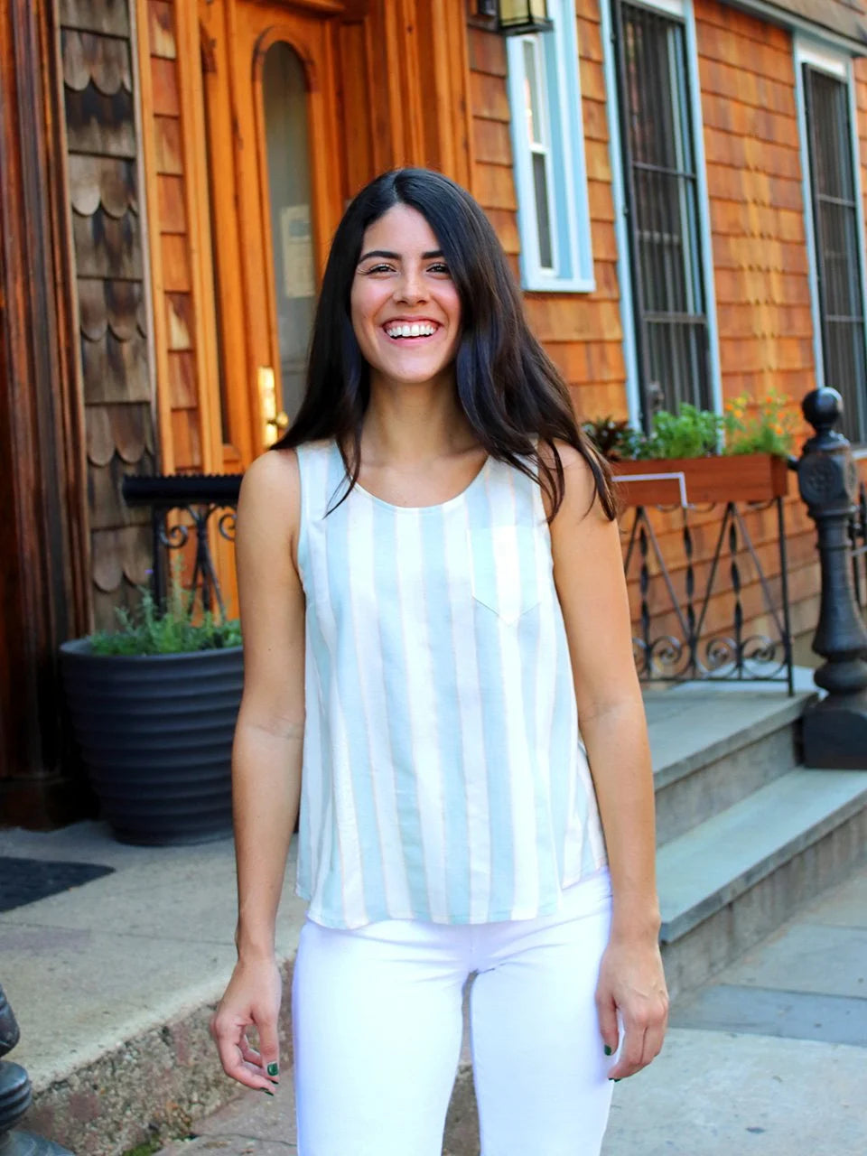 Woman in a white sleeveless top and pants standing in front of a wooden house.