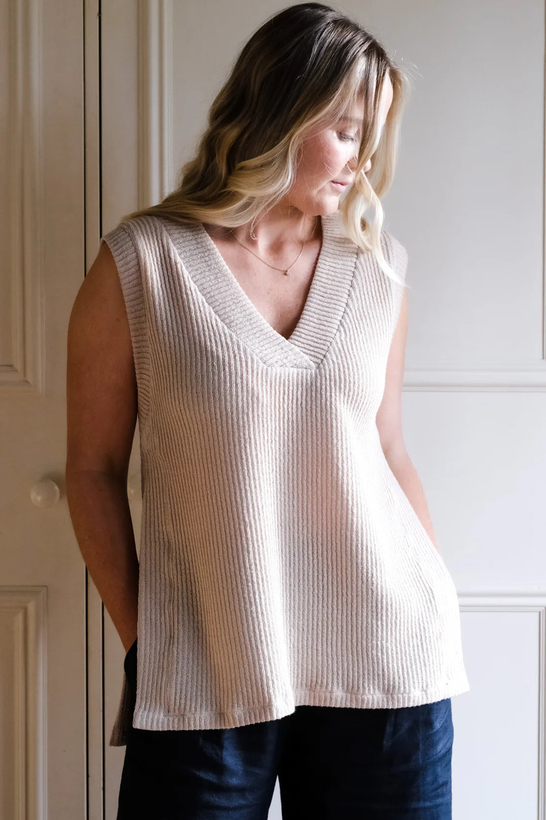 Woman wearing a sleeveless beige top and blue jeans against a white wall.