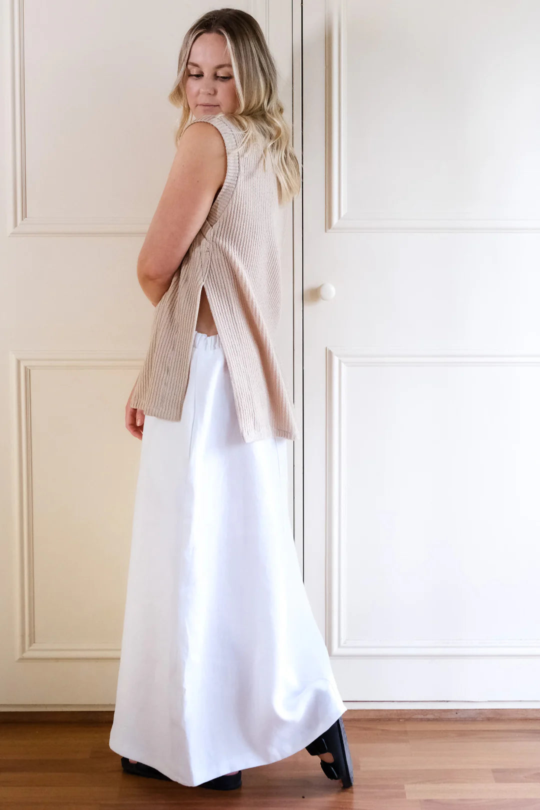 Woman wearing a beige sleeveless sweater over a white dress in a room with white walls and wooden floor.