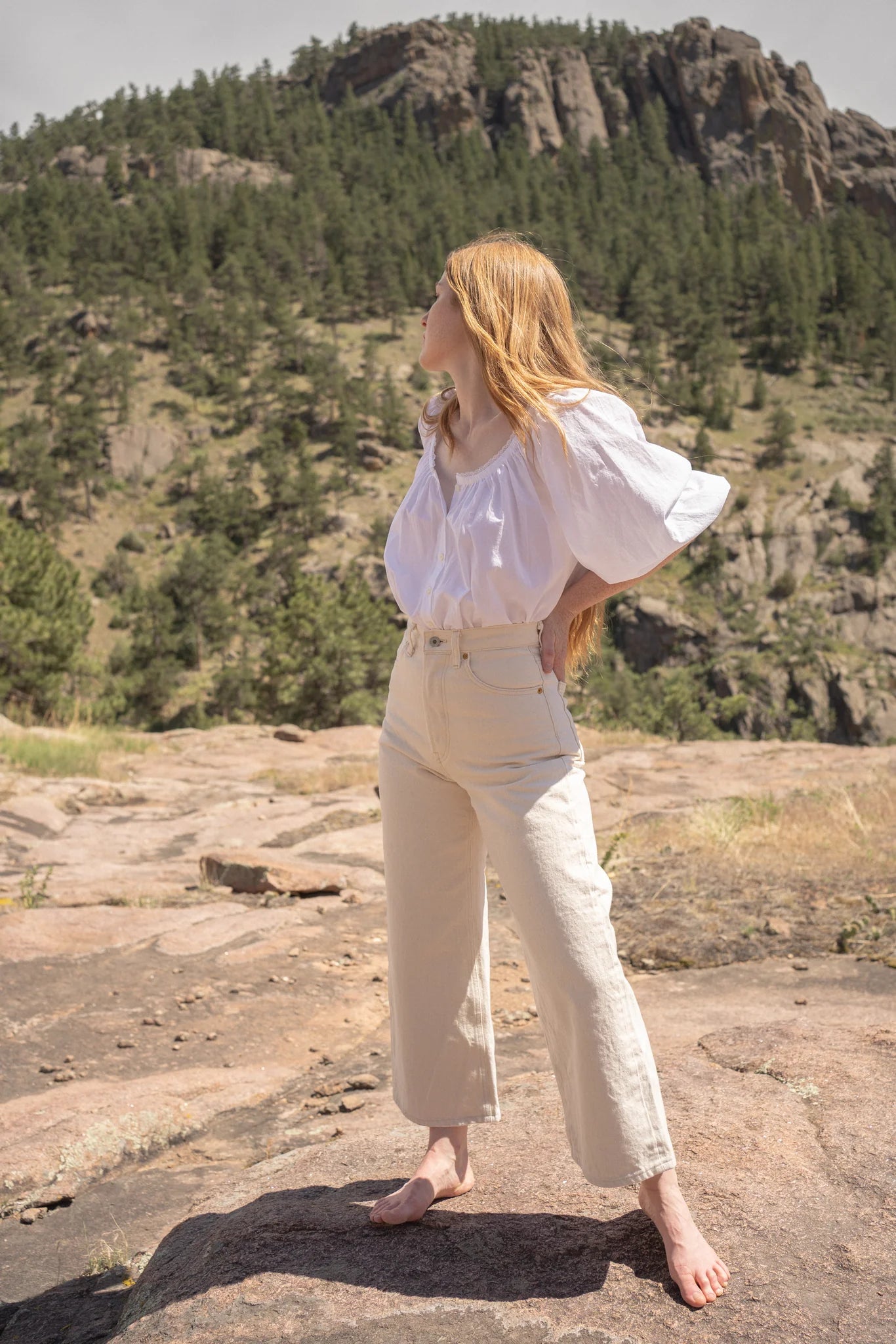 Woman in a white blouse and beige pants standing on a rocky outcrop with a mountainous background